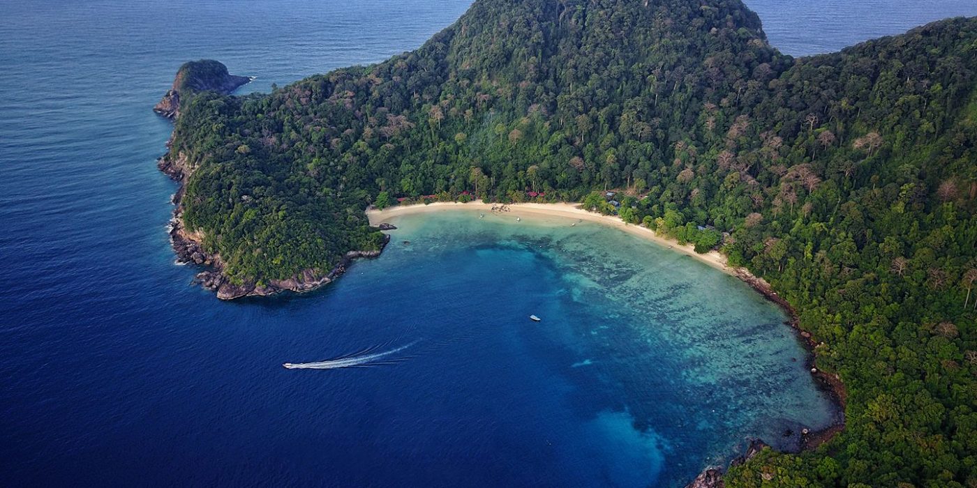 Aerial view of Tenggol Island crescent bay with a speedboat, white sand beach, and dense jungle hillside